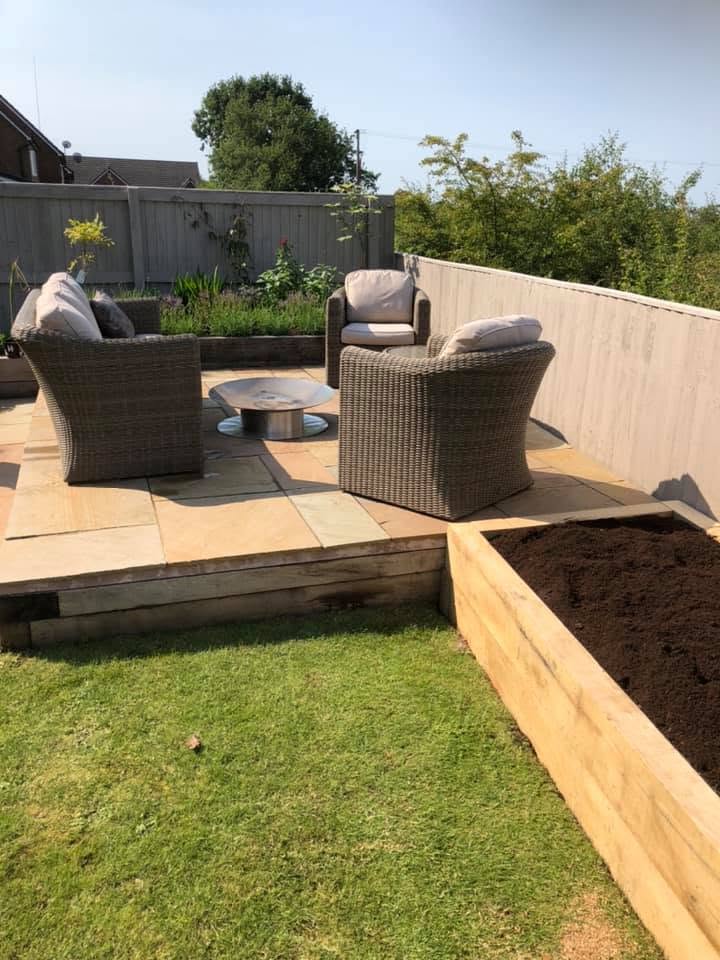 Raised sandstone patio seating area with rattan furniture and oak sleeper raised beds in bright sunshine