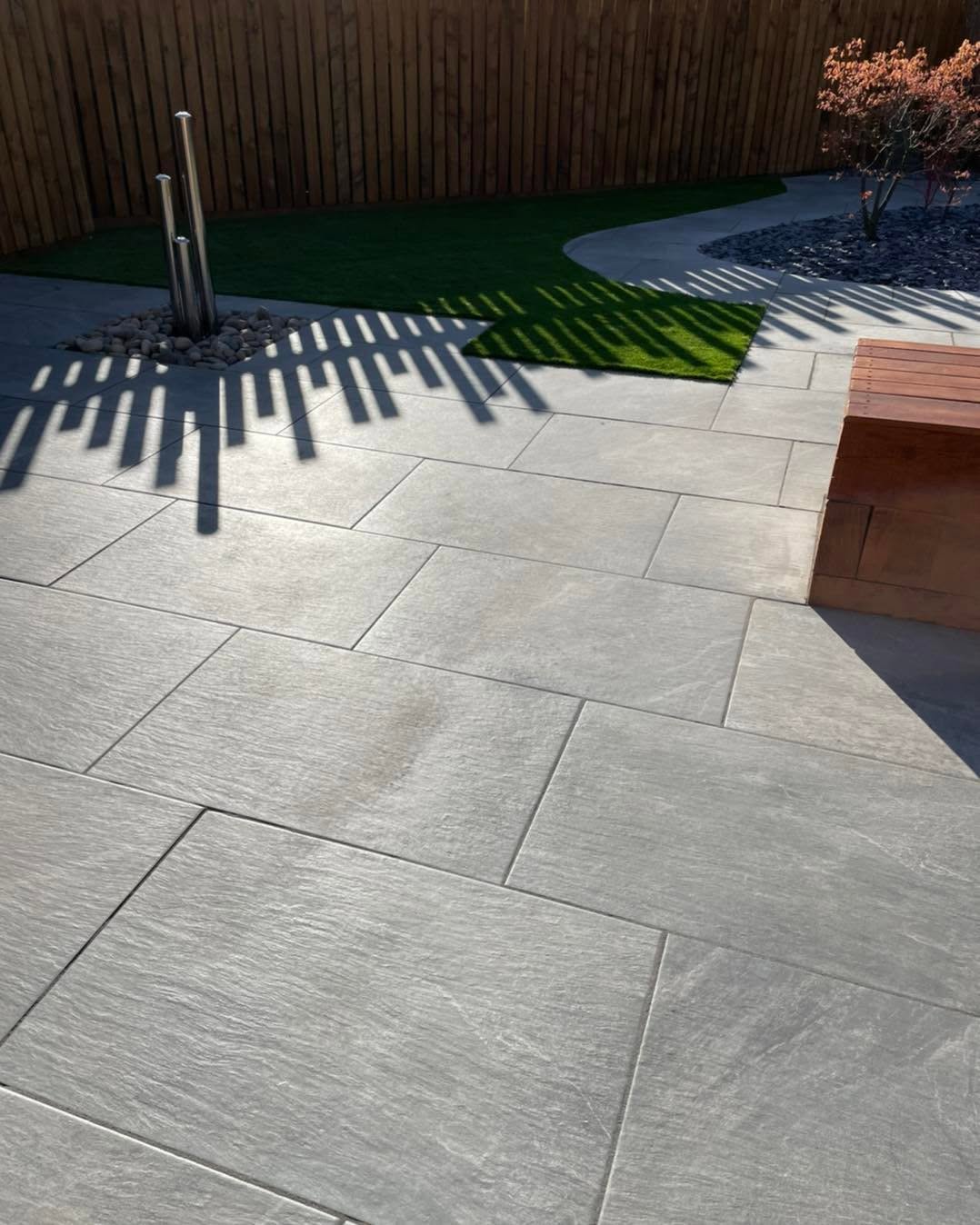Dramatic grey porcelain patio with stripe shadows from fence slats, artificial grass and raised cedar bench